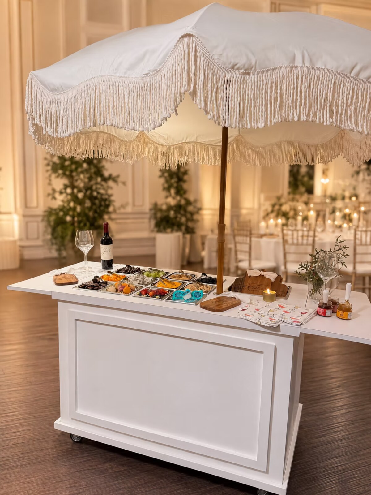 The Graze Carté cart: a white serving cart dressed with a fringed umbrella, laid out with cured meats, cheese, fruit and sweets at an evening event